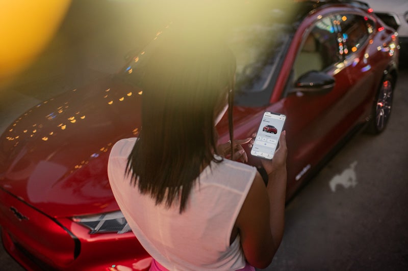 Woman using the FordPass® App on a phone while standing beside a Ford Mustang Mach-E® SUV 